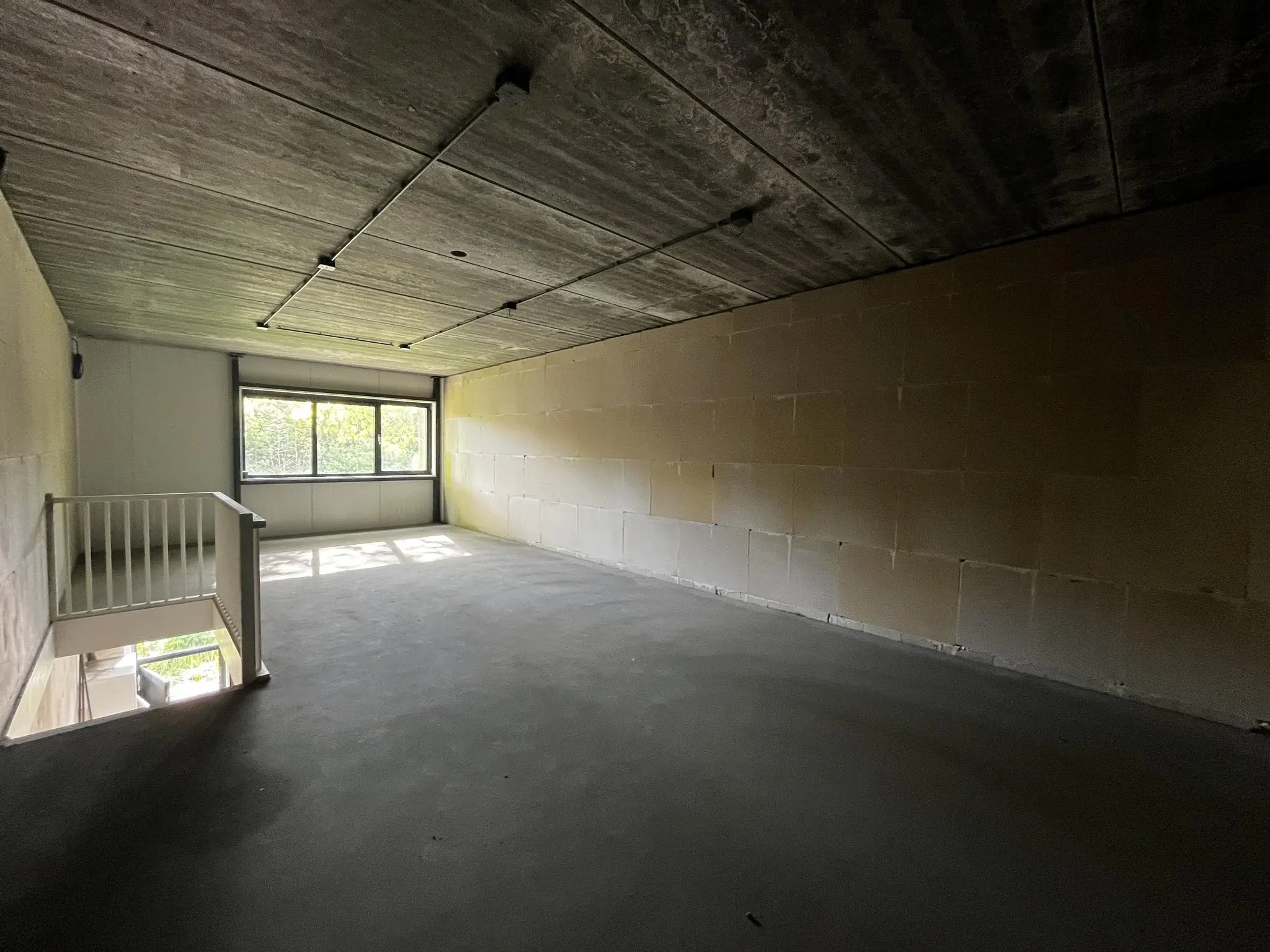 Lege ruimte met betonnen vloer en plafond, groot raam aan het einde en trapgat met witte leuning links.